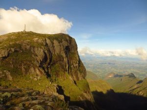 Audiodescrição: foto em dia ensolarado do Pico da Bandeira, um rochedo que parece alcançar o céu, devido às nuvens próximas a ele. Fim da audiodescrição.