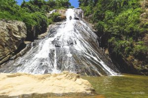 Audiodescrição: foto em dia ensolarado da Cachoeira das Andorinhas em Santa Leopoldina. A água escoa de um rochedo rodeado por mata verde. Fim da audiodescrição.