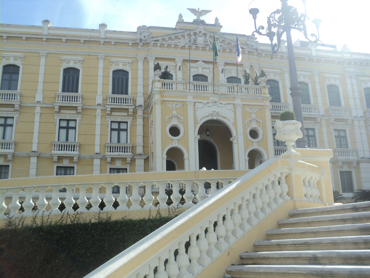 Audiodescrição: foto em dia ensolarado do Palácio Anchieta, visto de frente a partir da escadaria. A construção é amarela com detalhes brancos. Fim da audiodescrição.
